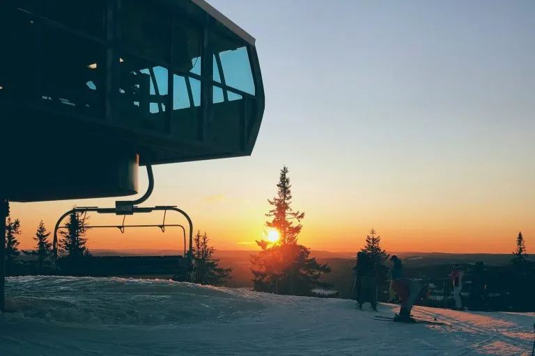 Slēpošanas trase ar modernām ēkām un saulrietu aiz tālām kokiem.