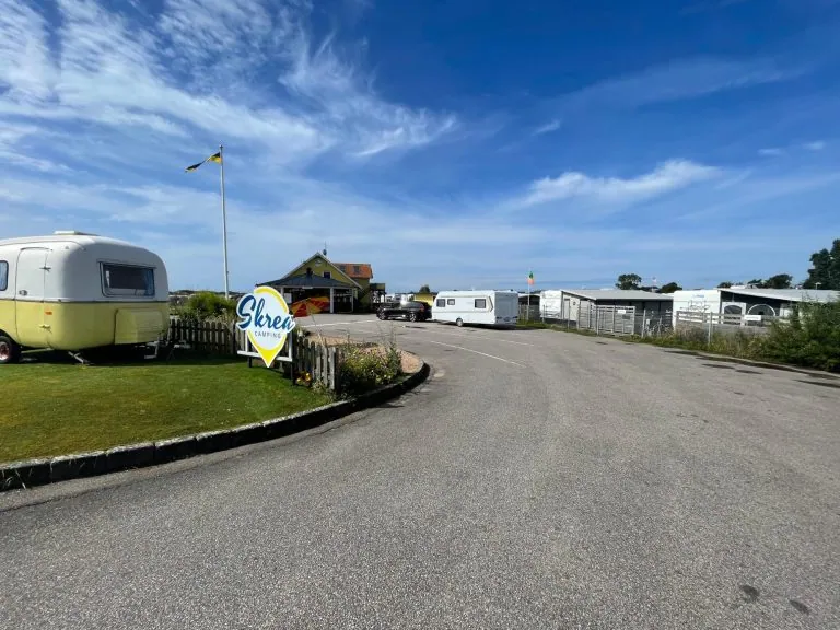 En campingplads med campingvogne og en bygning, omgivet af grønne områder og blå himmel.