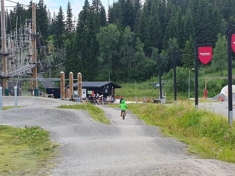 En cyklist i en grøn trøje på en grusvej, omgivet af træer og bygninger.