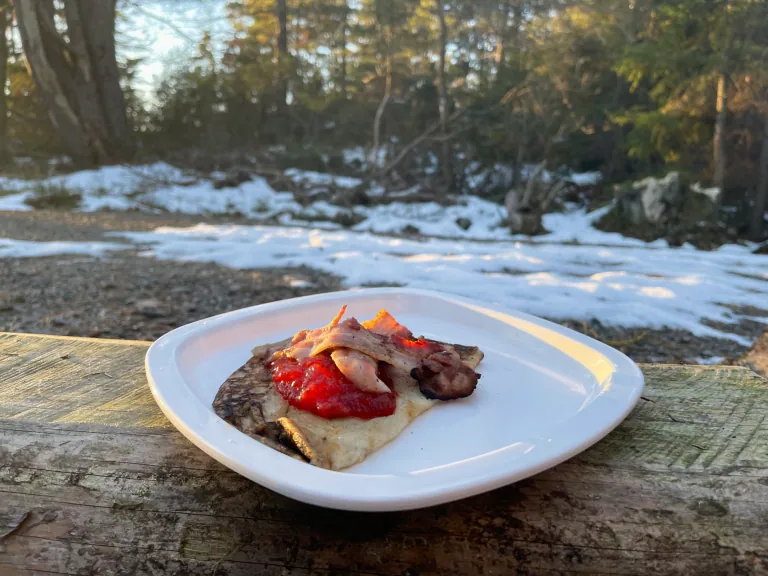 Falukorv burger with egg, mustard, and ketchup on a bench, surrounded by snow and nature.