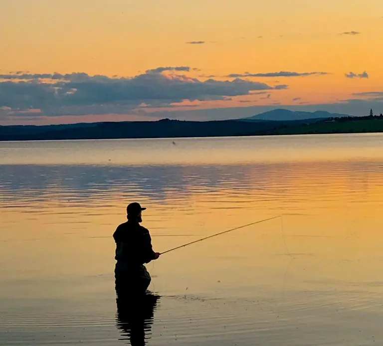 A person stands in a calm lake with fishing equipment against the sunset.