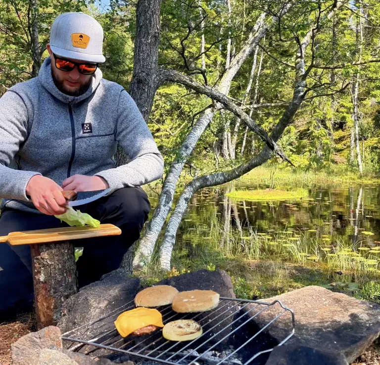 Henkilö grillaamassa purilaisia puiden ympäröimän järven rannalla.