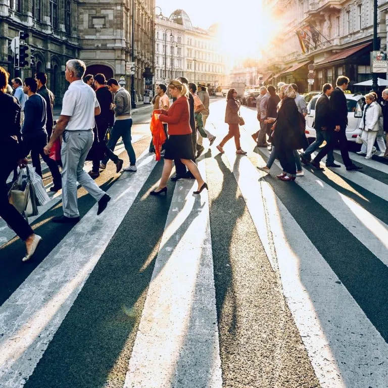 Folk går over ett övergångsställe i en stad med solnedgång i bakgrunden.