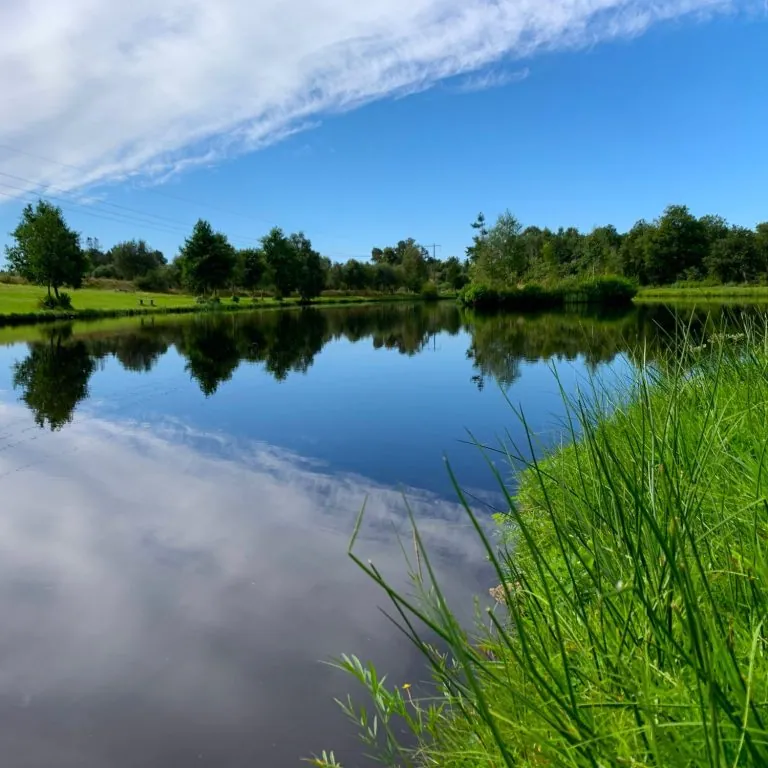 Het spiegelgladde meer omgeven door groene velden en bomen onder een heldere blauwe lucht.