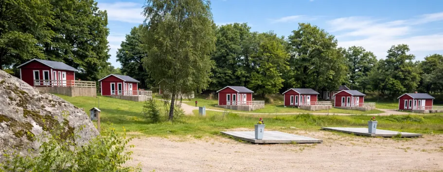 Røde hytter omgivet af grønne områder og træer, med sandet grund og moler i forgrunden.