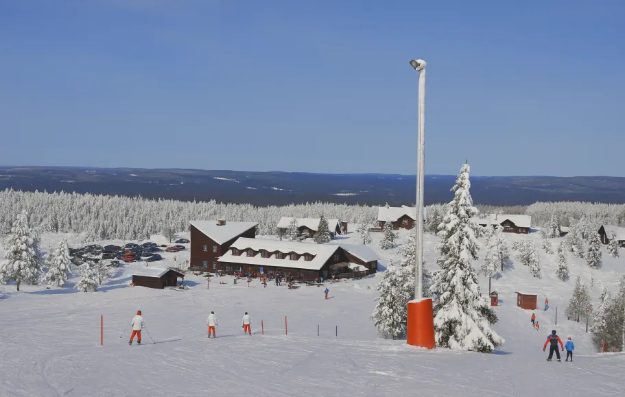 Snødekket skibakke med hytter og furutrær i bakgrunnen under en klar blå himmel.