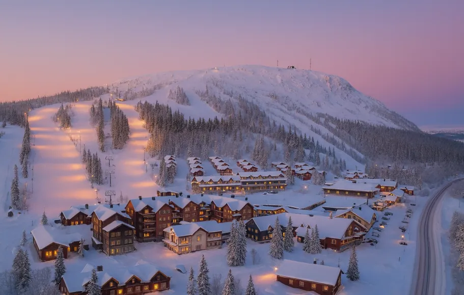 Talvimaiseman kuvitus lumipeitteisine mökkeineen ja laskettelurinteen auringonnousun värein.