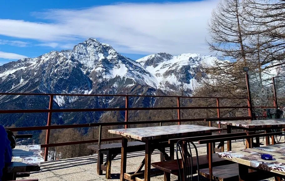 Blick auf schneebedeckte Alpenberge von einer Terrasse mit Holztischen und Bänken.