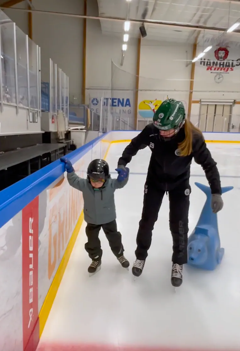 Felizia Wikner hilft Leo beim Eislaufen in einer Eisbahn. Beide tragen Helme.