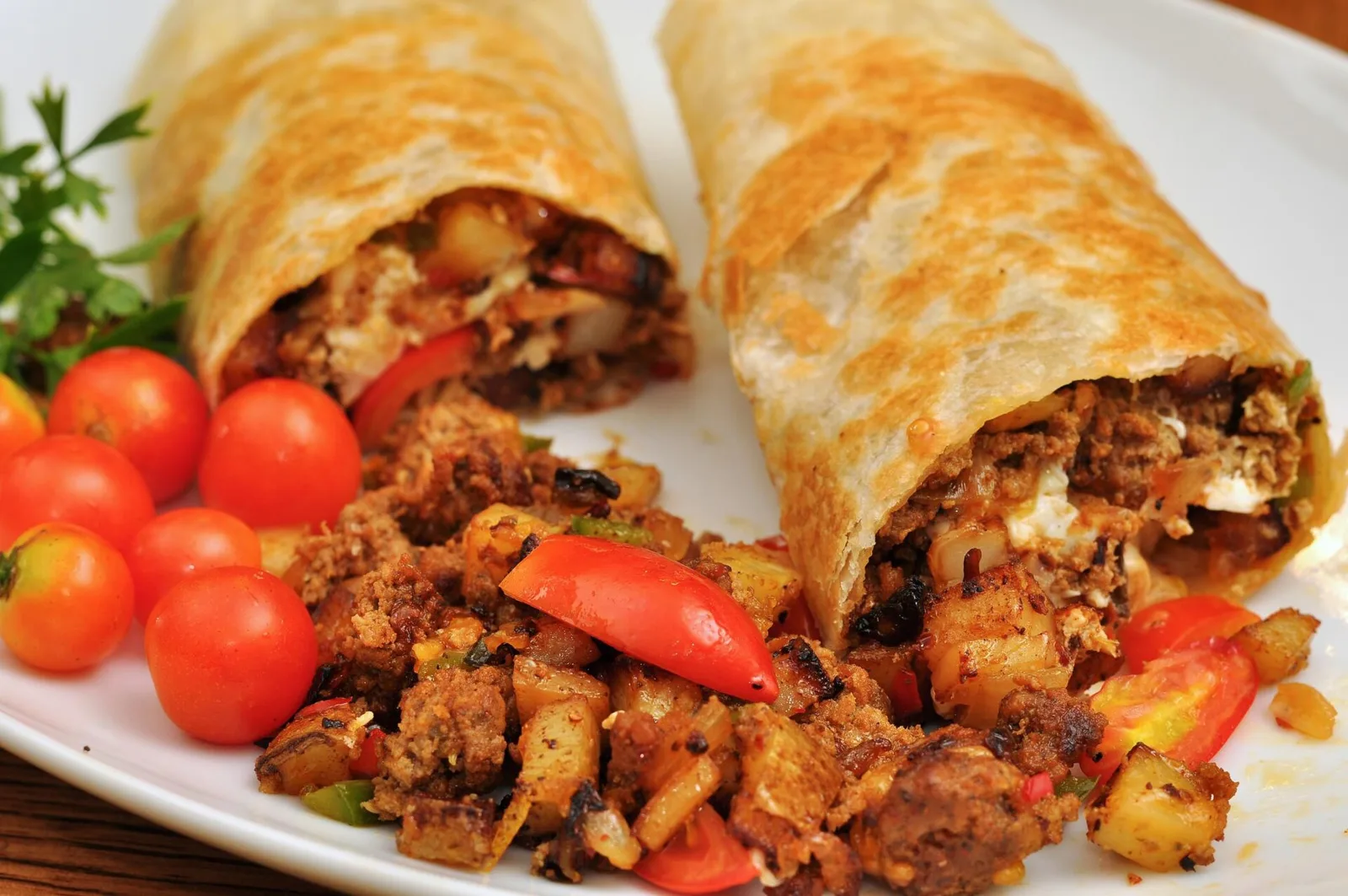 Två gyllene frukostburritos fyllda med ägg, potatis och kött på en tallrik med körsbärstomater.