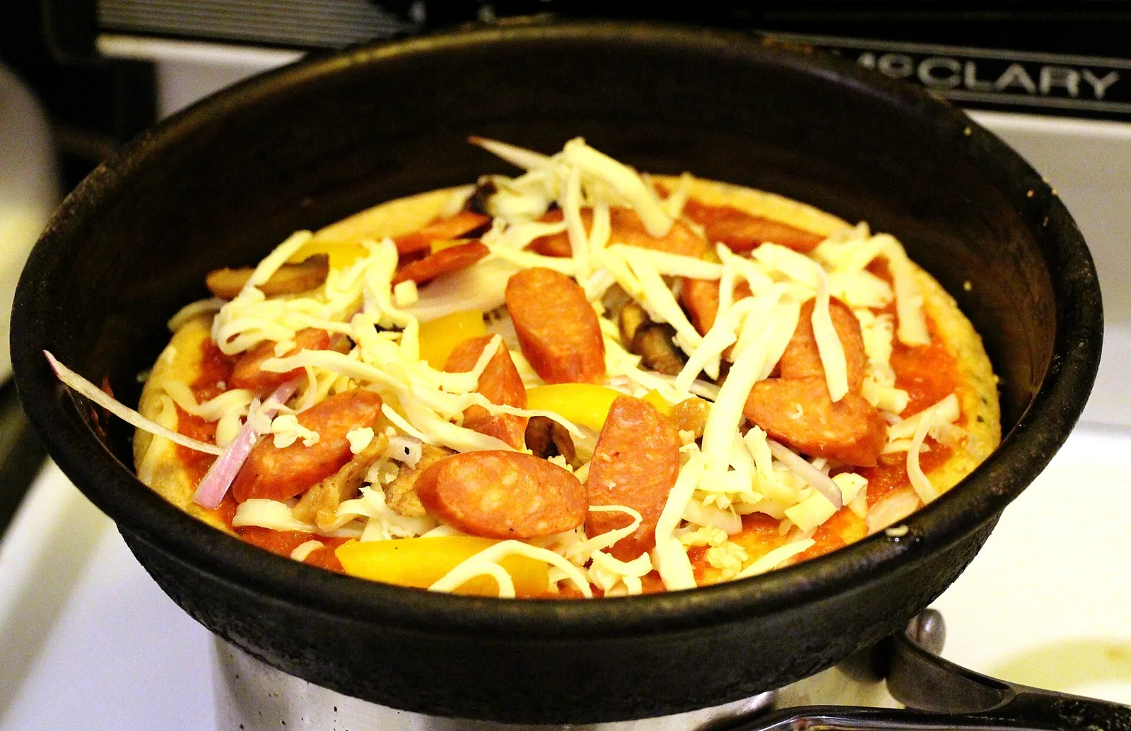 En ubagt pizza i en sort støbejernspande, toppet med tomatsauce, pølse, sød peber, løg og revet ost.