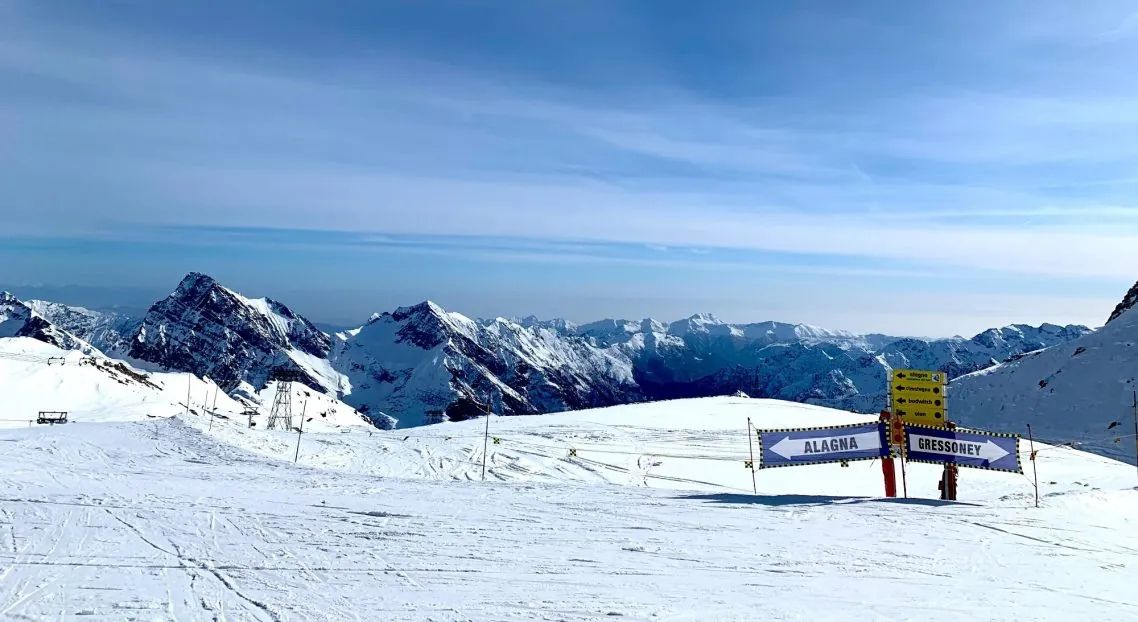 Slieksnis sniegā ar bultām uz Alagna kreisajā pusē un Gressoney labajā pusē, dzeltenās pistes zīmes un kalnu grēdas fonā