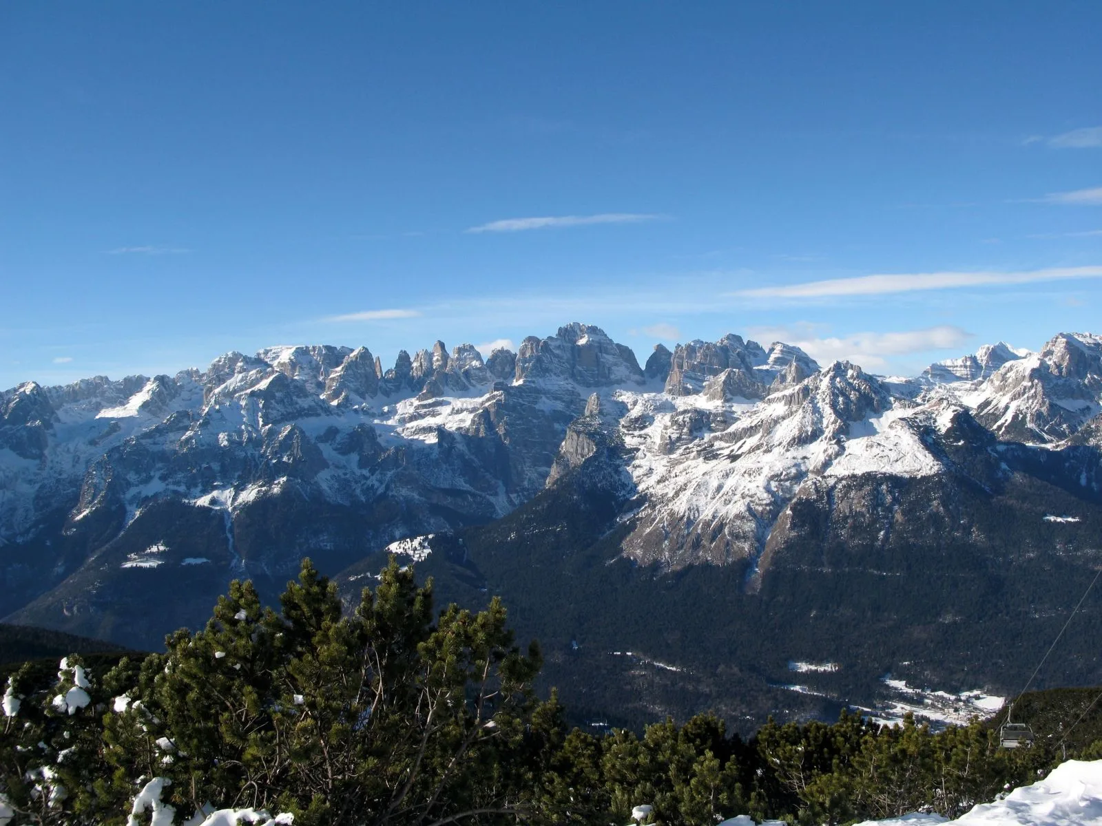 Udsigt fra Paganella en klar vinterdag mod Brenta-dolomitternes snedækkede, takkede tinder, med bjergfyr og en stolelift i forgrunden