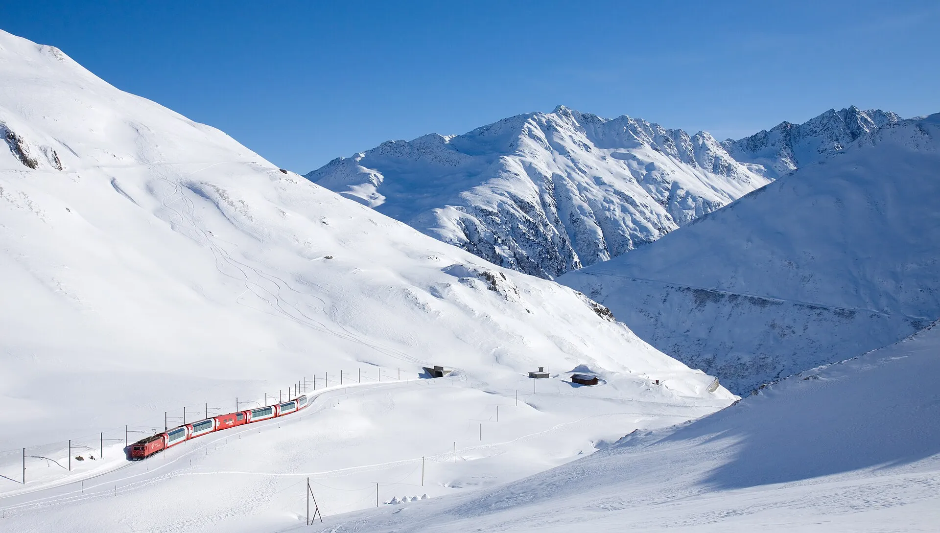 Ett rött tåg kör genom ett djupt snölandskap mot Oberalppasset ovanför Andermatt, med snötäckta toppar under en klarblå himmel.