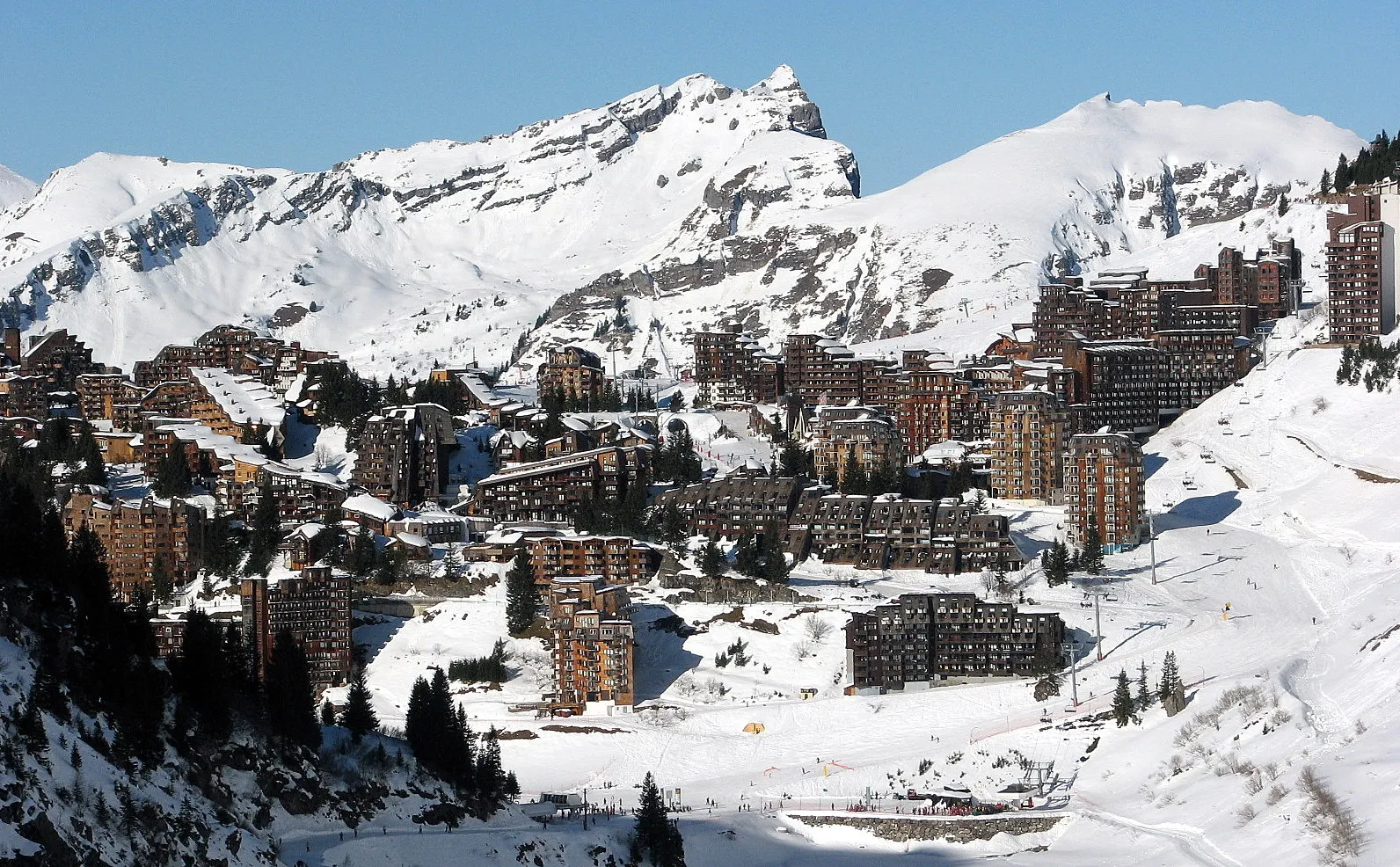 Den träklädda skidbyn Avoriaz på en snöig bergssluttning under höga, snötäckta toppar och klarblå himmel