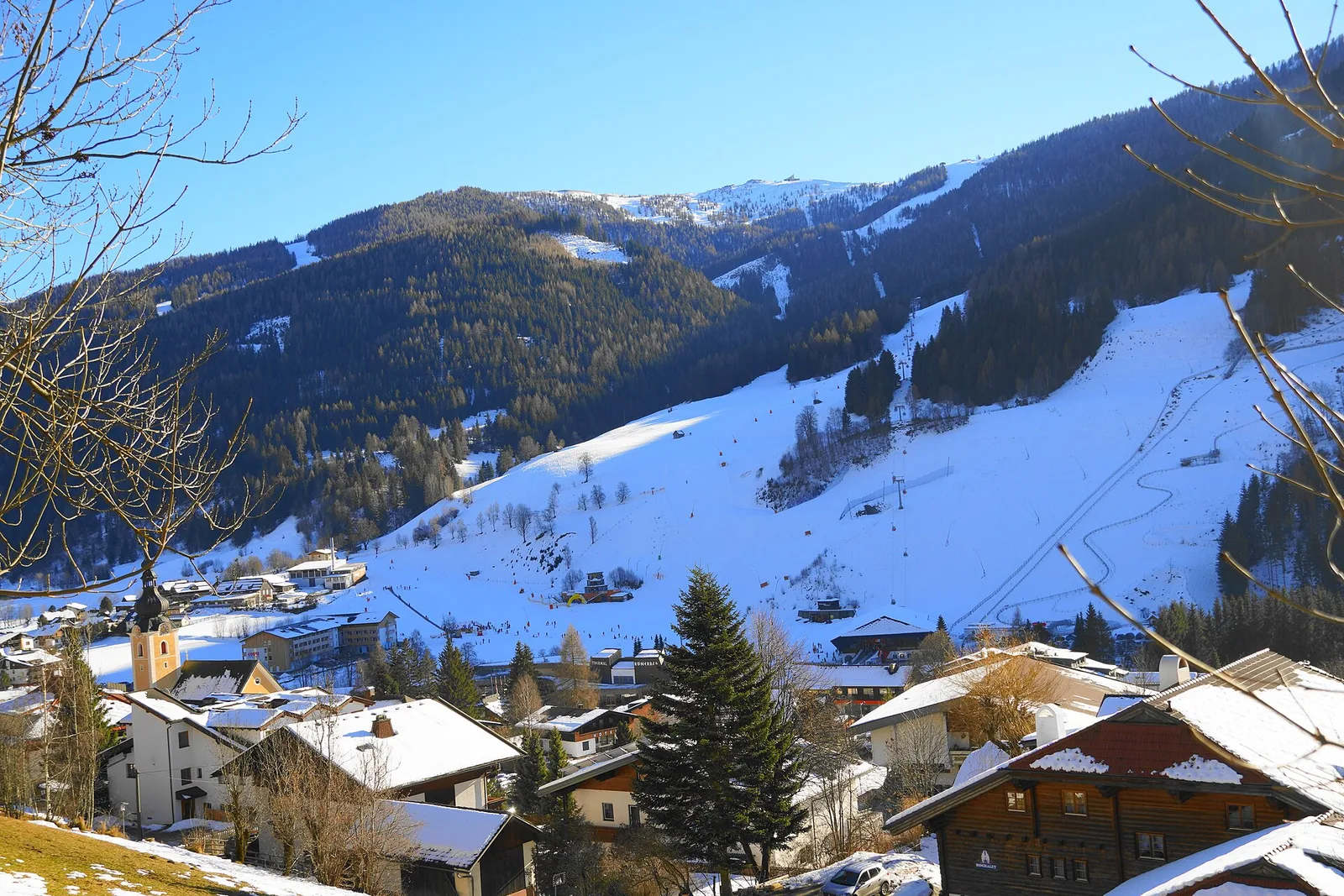 Bad Kleinkirchheim w słoneczny zimowy dzień, z wieżą kościoła i zaśnieżonymi dachami na pierwszym planie, Thermal Römerbad u podnóża stoku i trasami pnącymi się w stronę szczytu Kaiserburg, na których widać małe kropki narciarzy