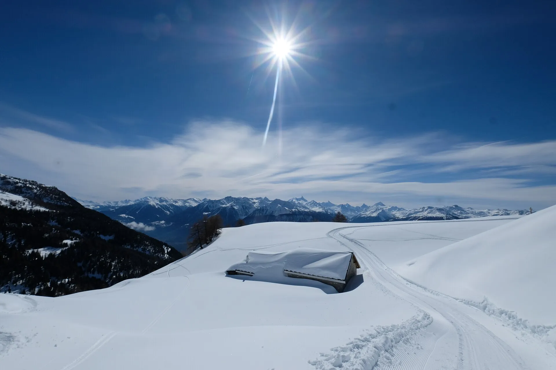 En insnöad stuga på en sluttning ovanför Crans-Montana i strålande sol, med snötäckta berg på andra sidan Rhônedalen.