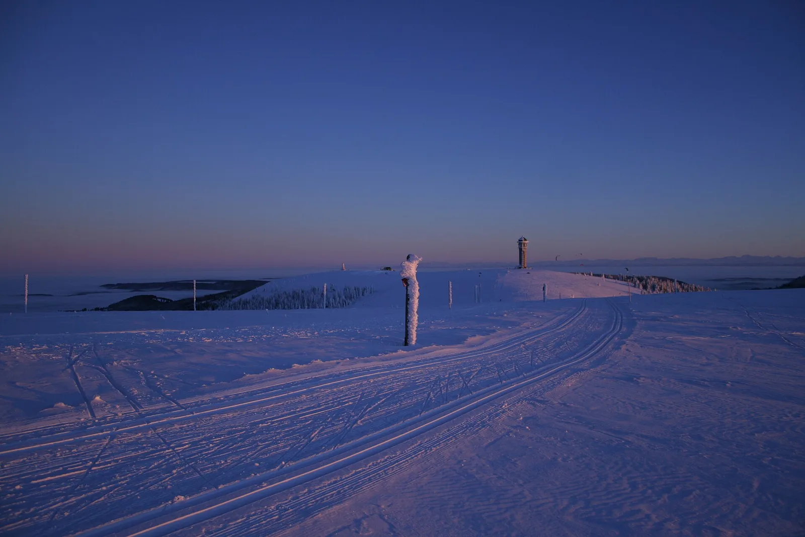 Nyspårat längdspår på Feldbergs kala topp i kvällsljus, med Feldbergturm på Seebuck och Alperna vid horisonten ovanför ett molnhav.