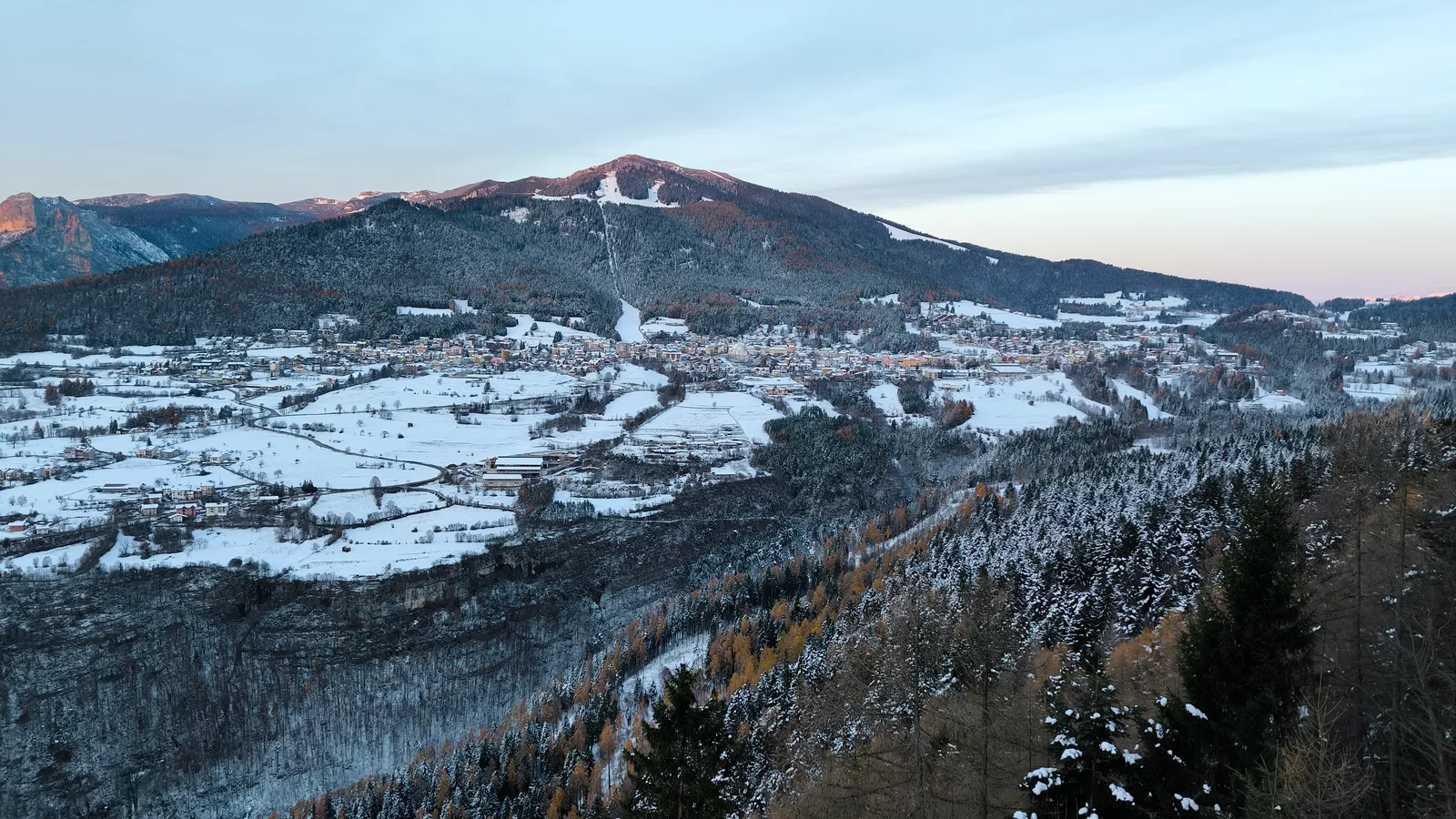 Snötäckta ängar och byn Folgaria i skymningsljus, med pisterna nedför Monte Cornetto i bakgrunden, sett från Serrada