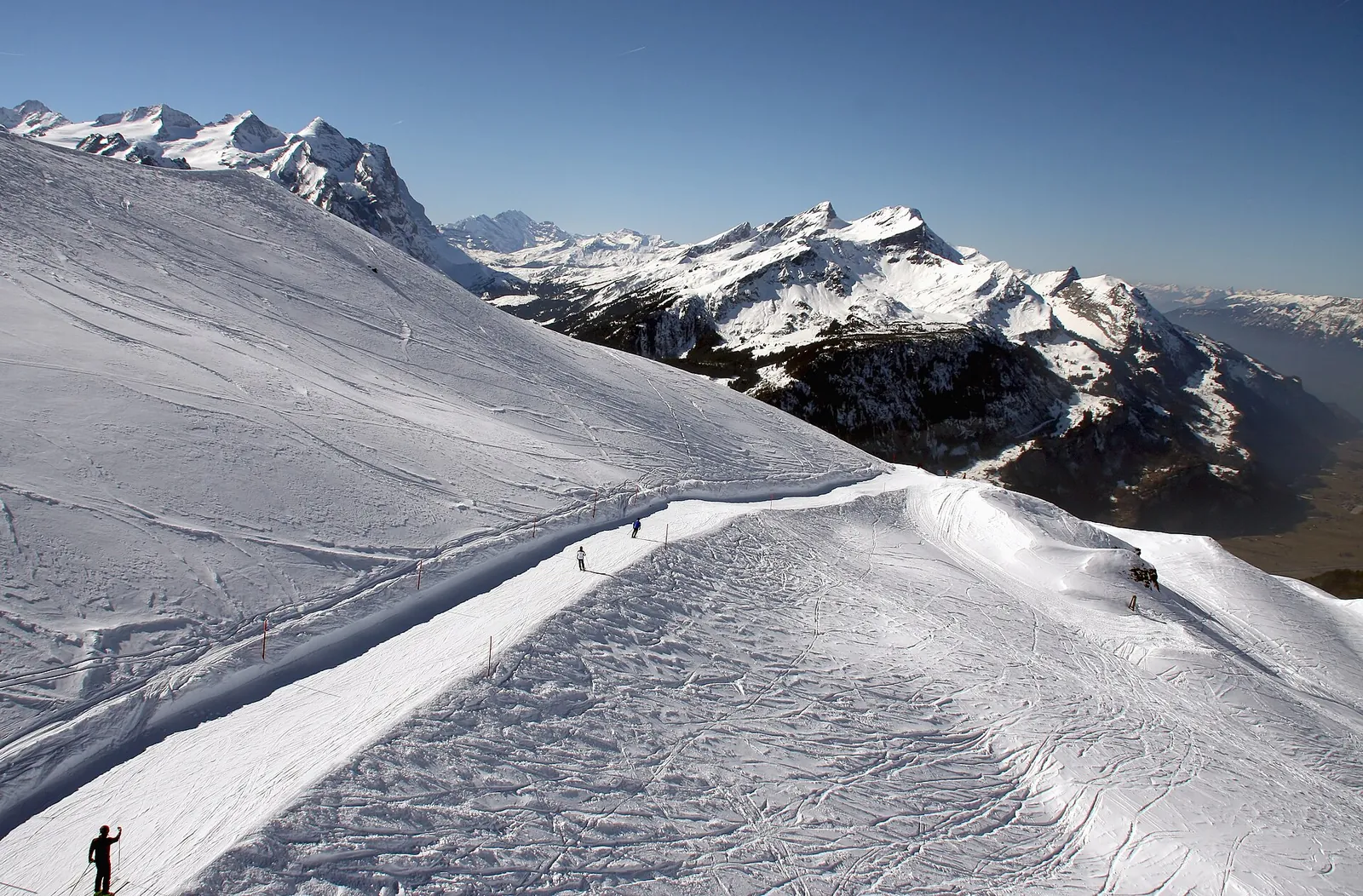 The piste from Planplatten towards Mägisalp on a sunny spring day, with a few skiers in the distance and snow-covered peaks of the Bernese Oberland in the background