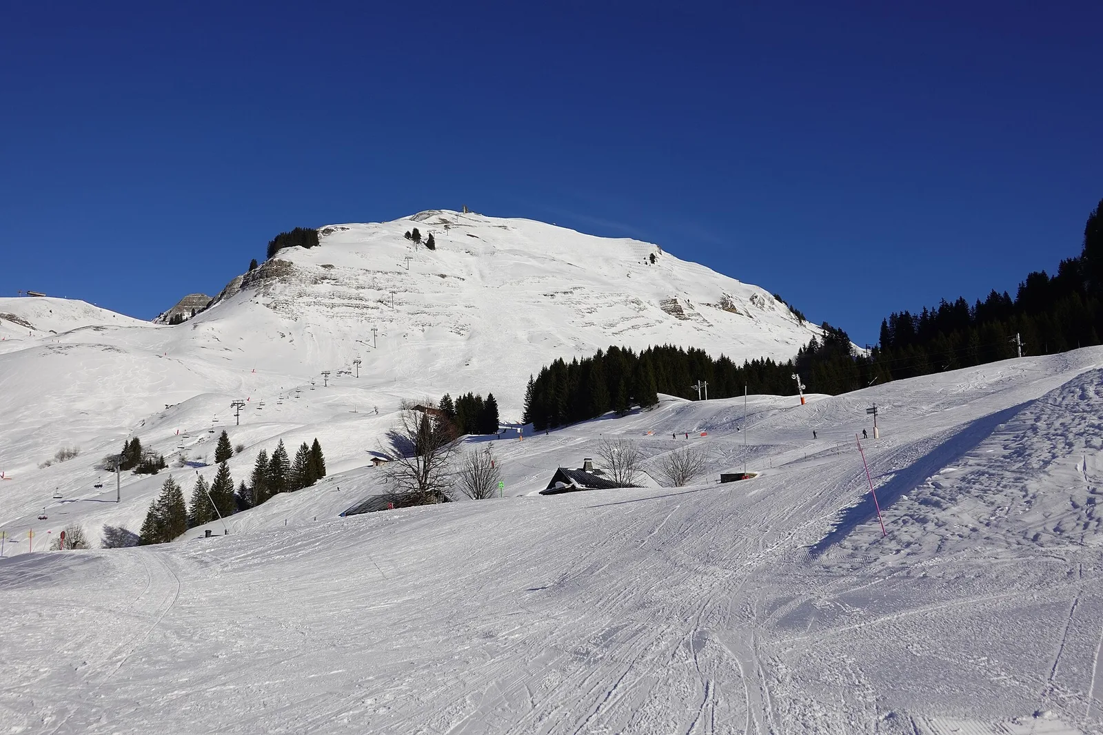 Широкі підготовлені траси в Le Grand-Bornand під засніженою горою з крісельними підйомниками, ялиновим лісом і старою фермою в сонячному світлі