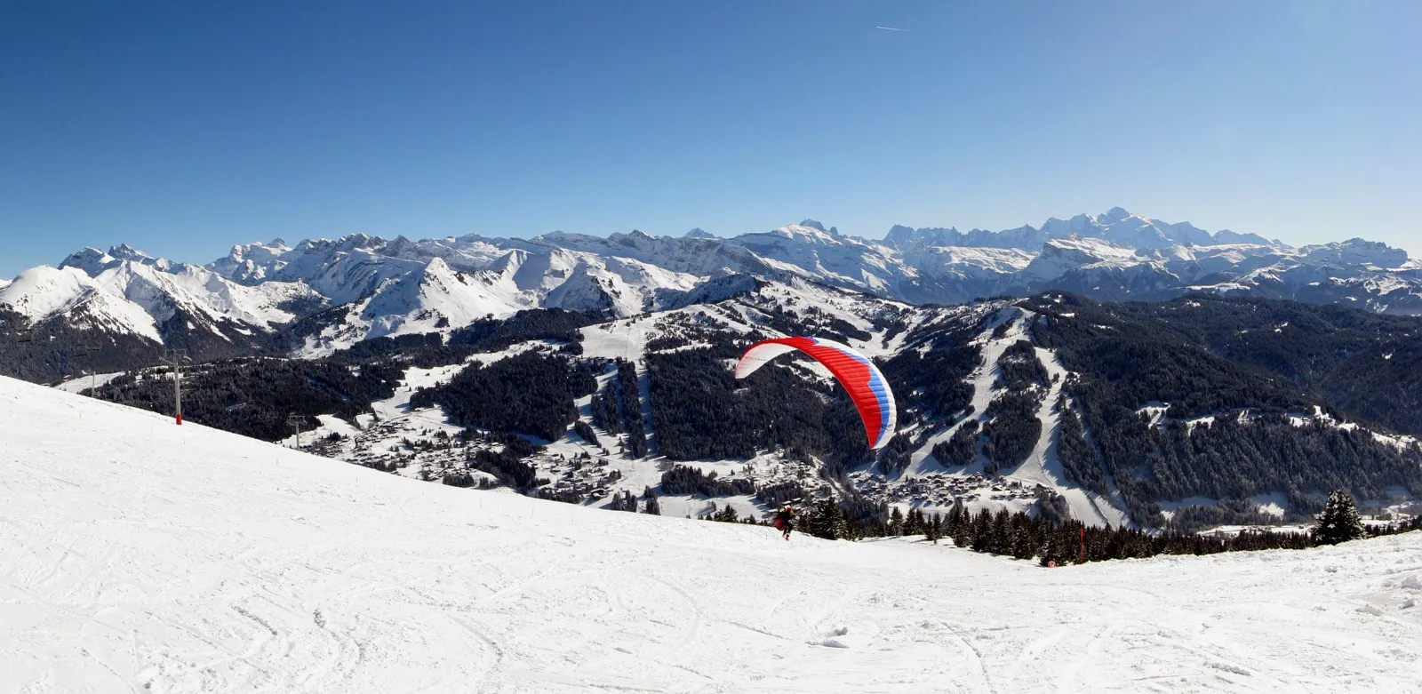 Utsikt från Mont Chéry över de snötäckta pisterna i Les Gets, med en skärmflygare i luften och Mont Blanc-massivet vid horisonten.