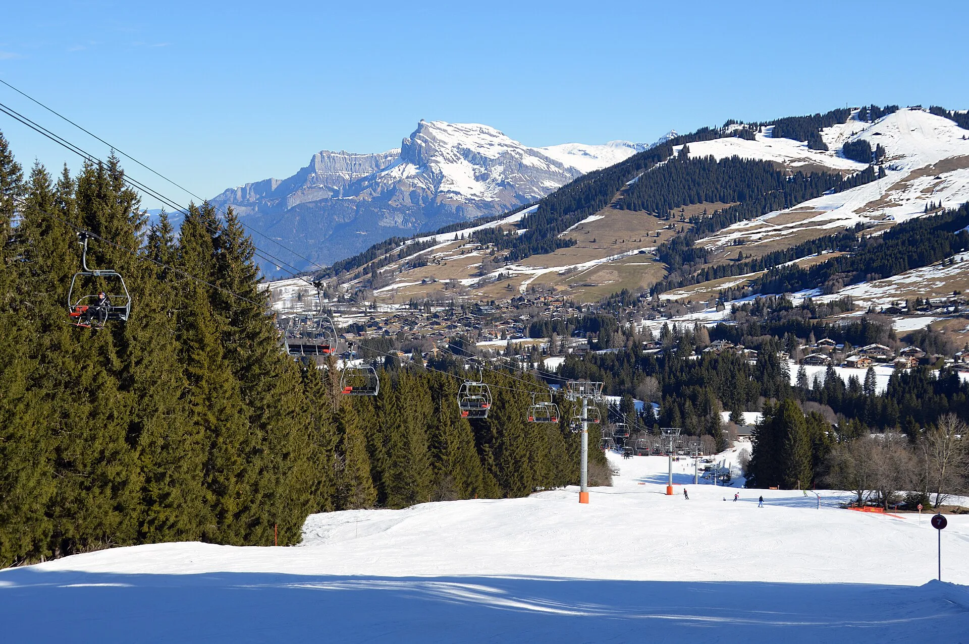 Utsikt nerför en pist i Megève mot byn i dalen, med en stolslift genom granskogen och snötäckta berg i bakgrunden.