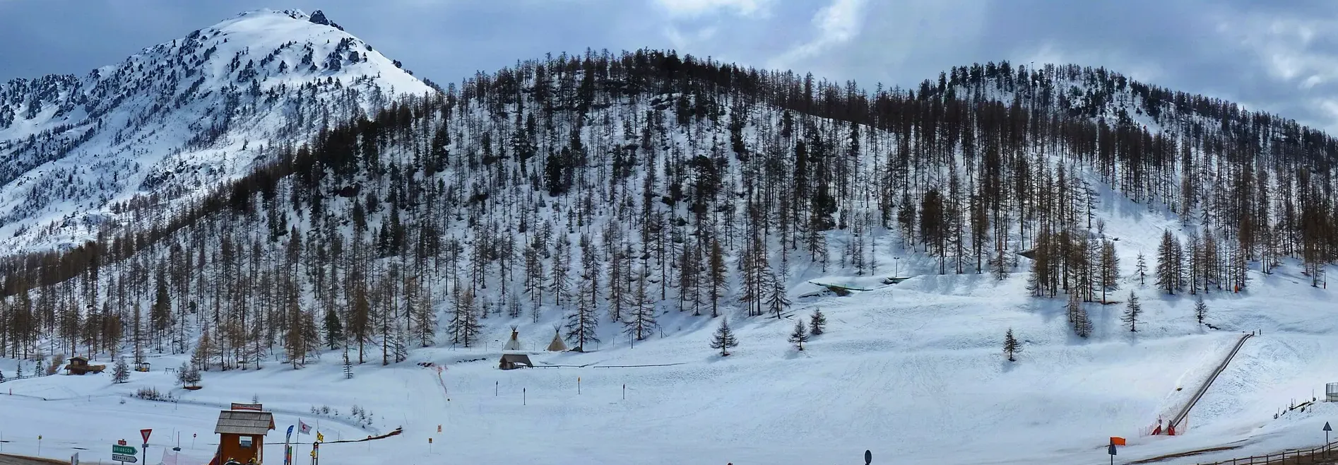 Nybörjarbacken mitt i Montgenèvre med rullband och tipitält, nedanför en snötäckt sluttning med lärkskog och en spetsig topp.