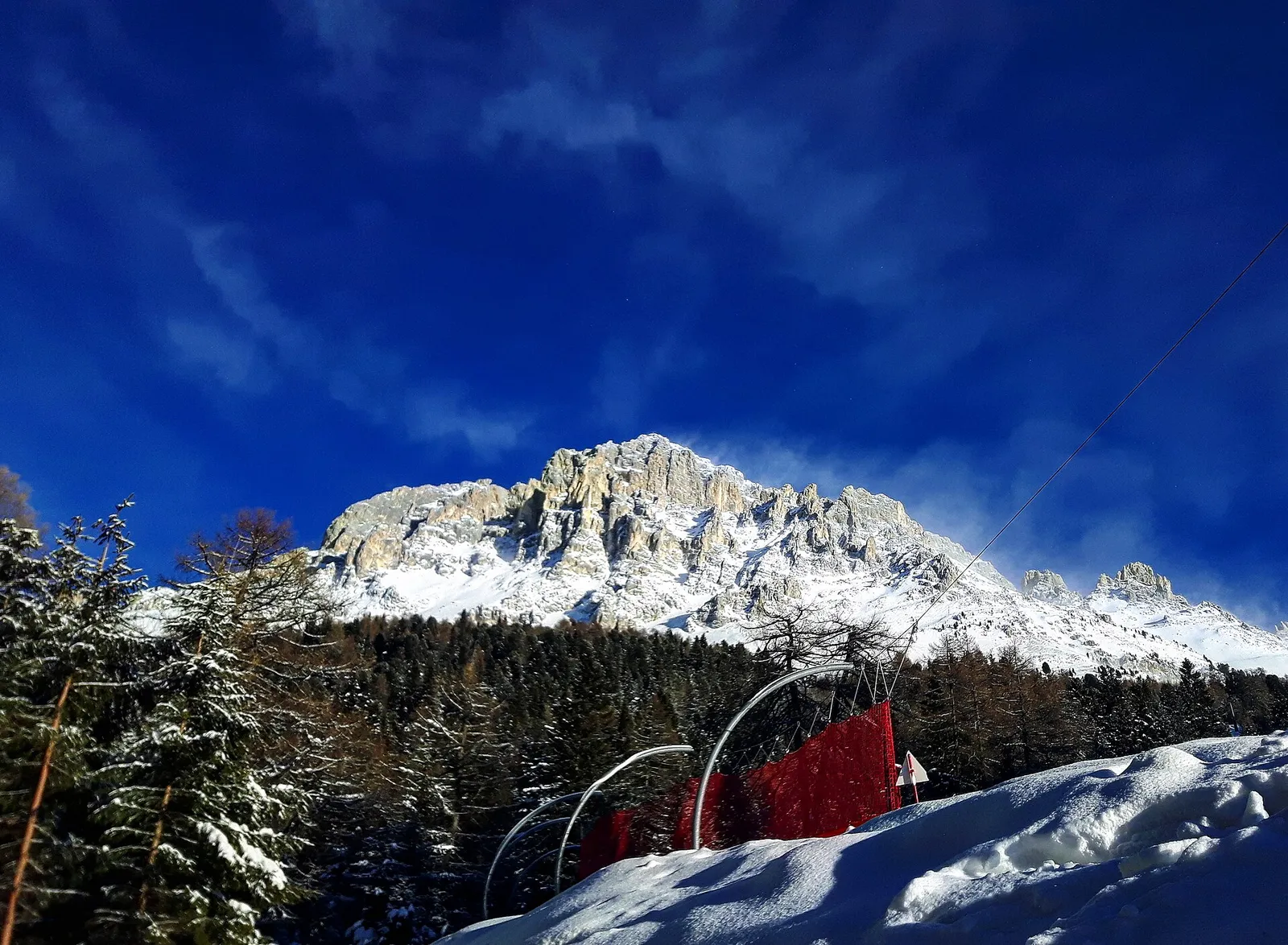 Verschneite, zackige Dolomitengipfel vor tiefblauem Himmel über Fichtenwald und einem roten Fangnetz am Pistenrand in Obereggen