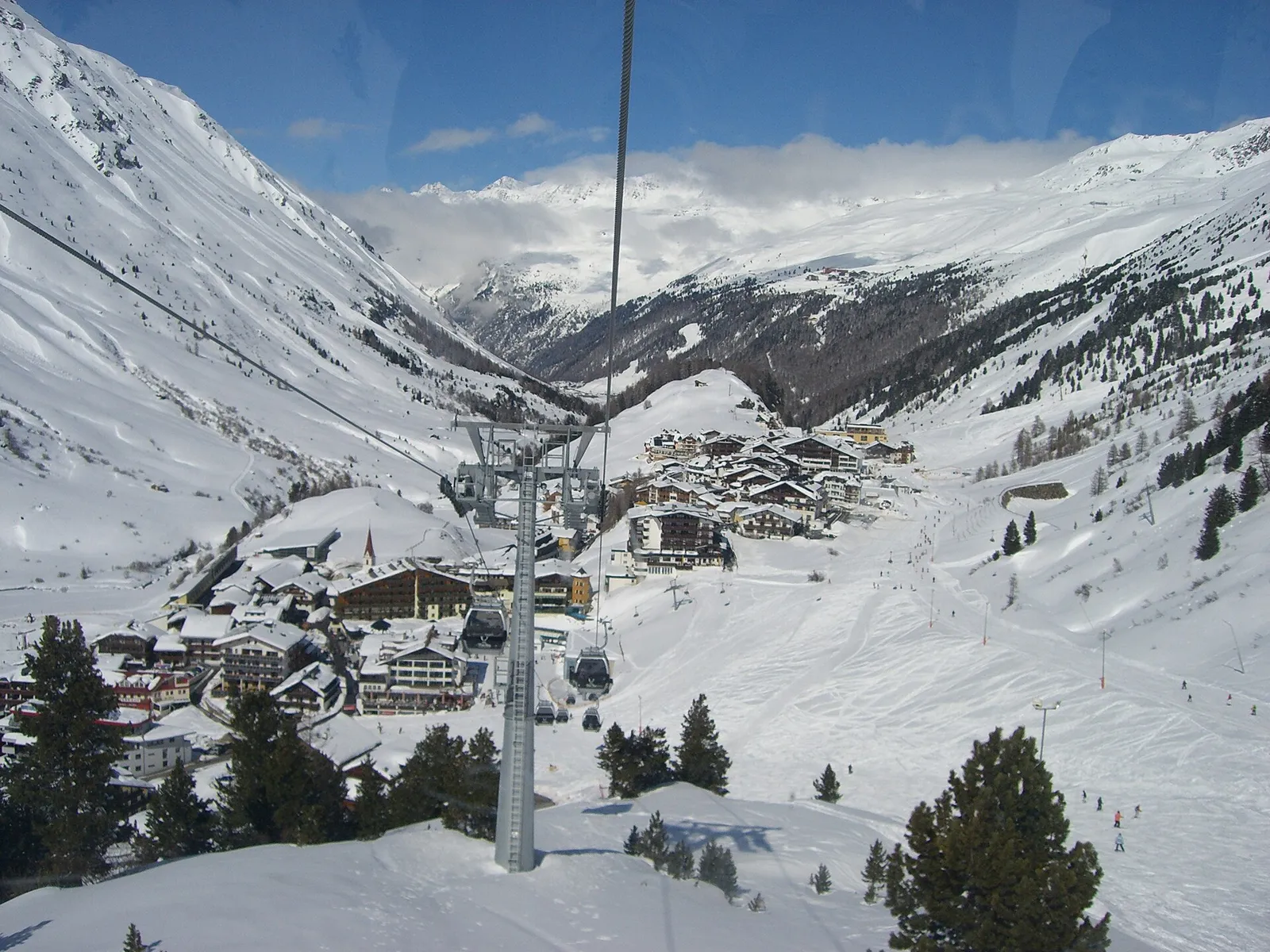Obergurgl im Winterkleid aus einer Gondel gesehen, mit den Hotels rund um die Kirche, breiten Pisten hinunter zum Dorf und verschneiten Bergen ganz hinten im Ötztal.