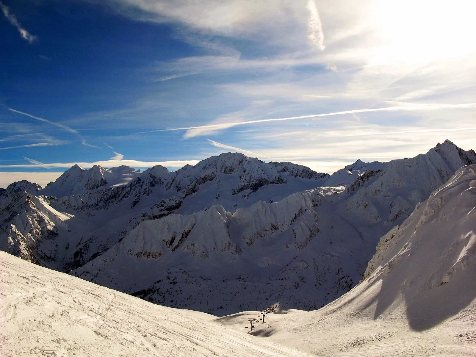 Ośnieżone szczyty masywów Presena i Presanella pod błękitnym niebem z mgiełką chmur, widziane z trasy Paradiso nad Passo Tonale, z wyciągiem krzesełkowym daleko w dolinie