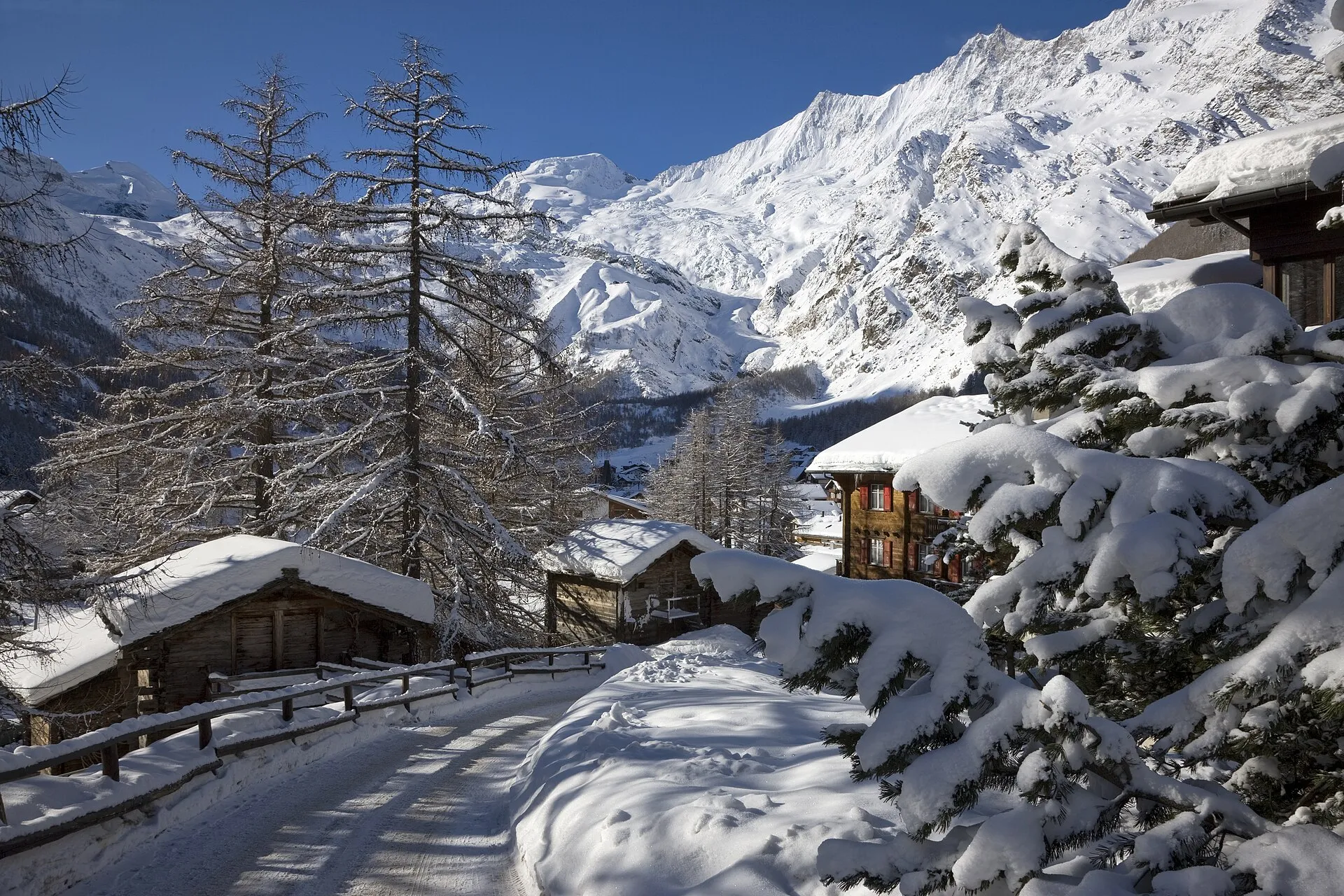 Den snötäckta byn Saas-Fee i en dalgång, omgiven av höga, glaciärklädda berg under en blå himmel.
