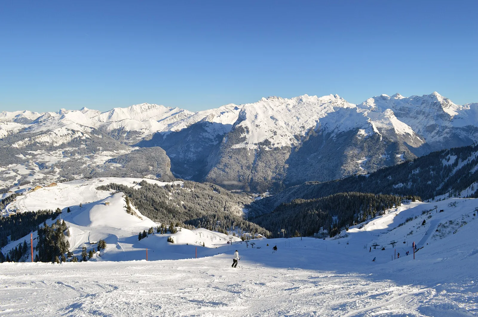 Bred pist nedanför Tête des Saix med utsikt mot snötäckta berg, granskog och platån Samoëns 1600 till vänster, med några skidåkare långt bort