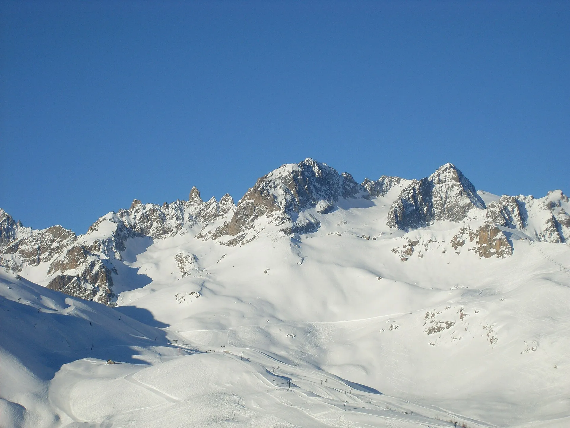 Taggiga, snötäckta bergstoppar ovanför de breda pisterna i Serre Chevalier under en klarblå himmel.