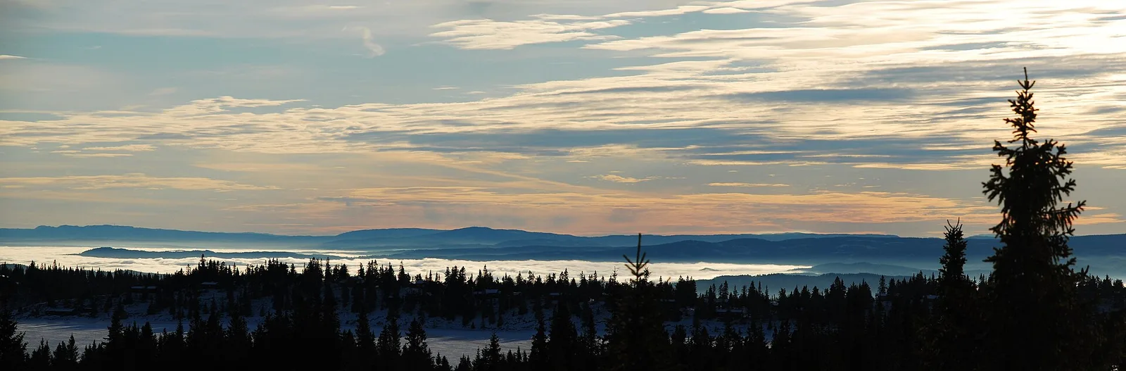 Utsikt från skogen vid Sjusjøen över dimmiga dalar och blå berg i låg kvällssol.