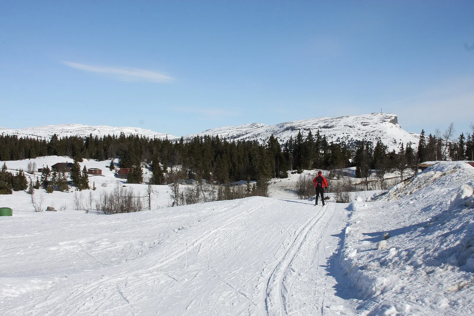 Ett preparerat längdspår i snön mot fjället Skeikampen med snötäckt topp, granskog och några stugor under klarblå himmel.