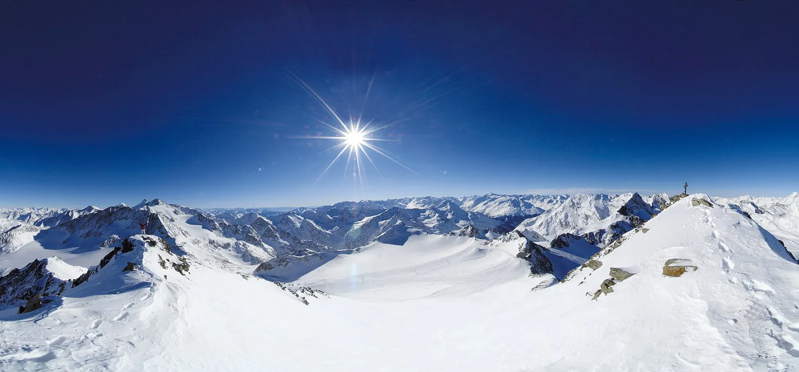 Vidvinkelpanorama från Schaufelspitze över Stubaier Gletscher, med snötäckta toppar, glaciärpister och en klarblå himmel med sol