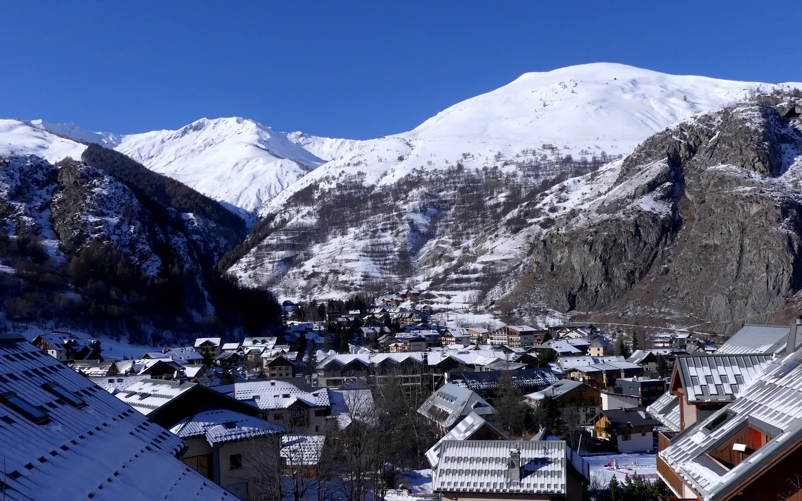 Verschneite Dächer in Valloire vor einem Tal mit weiteren Dorfhäusern, steilen Felswänden und weißen Berggipfeln unter klarblauem Himmel