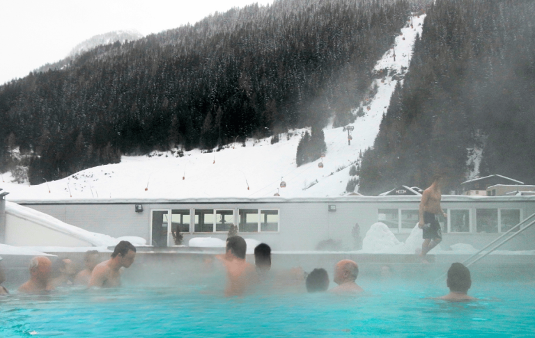 Personer badar i en varm källa omgiven av snö och alpberg. Dimmigt väder.