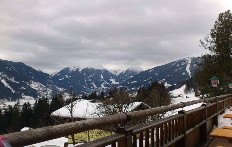 Utsikt över snötäckta berg och träd från en träplattform. Mörka moln på himlen.
