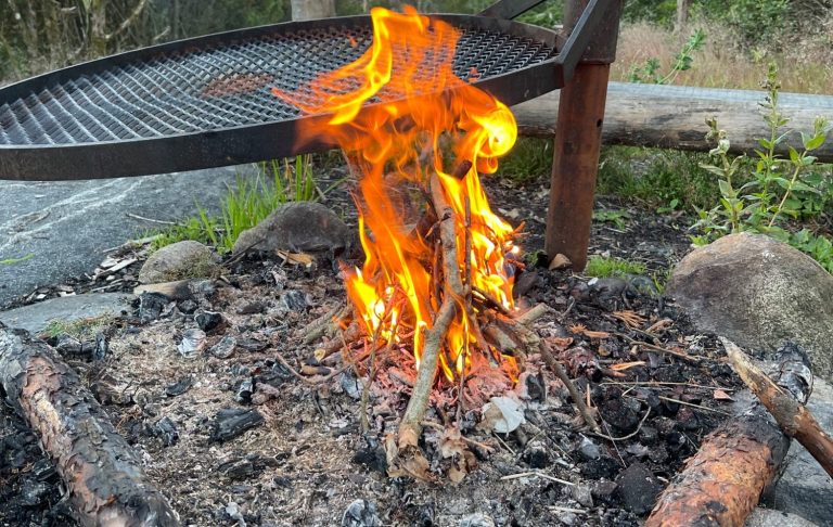 Eld som flammar över en ställning av grenar och aska vid en grillplats.
