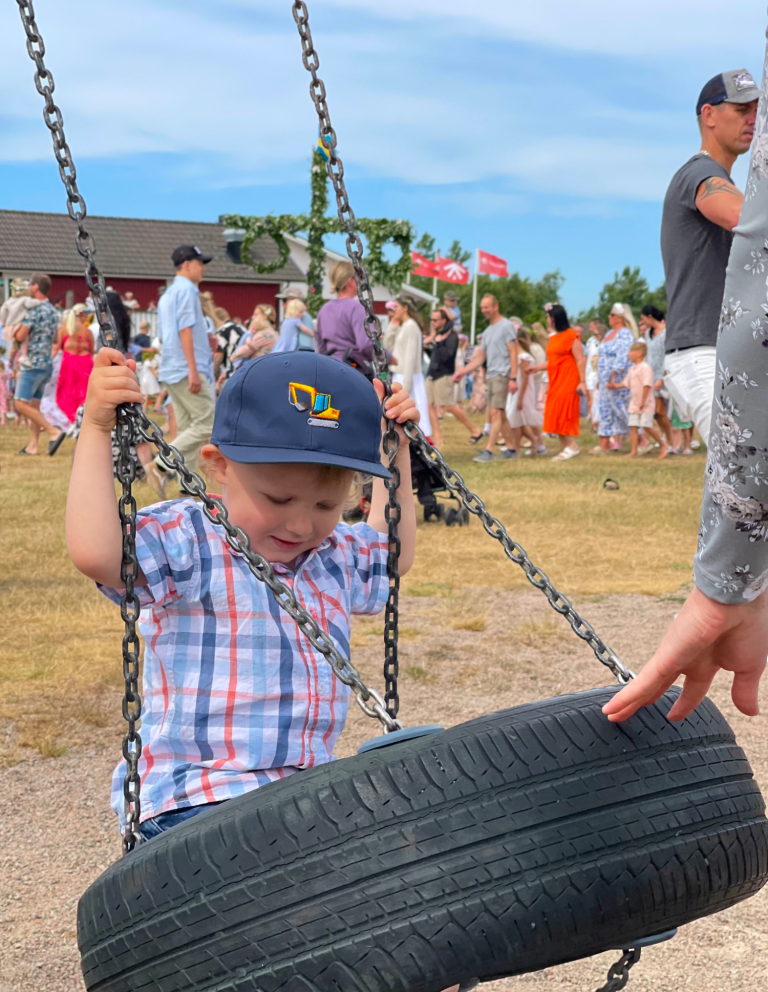 Litet barn i rutig skjorta sitter på en gungställning och leker med en däckgunga. Dans runt midsommarstången i bakgrunden.