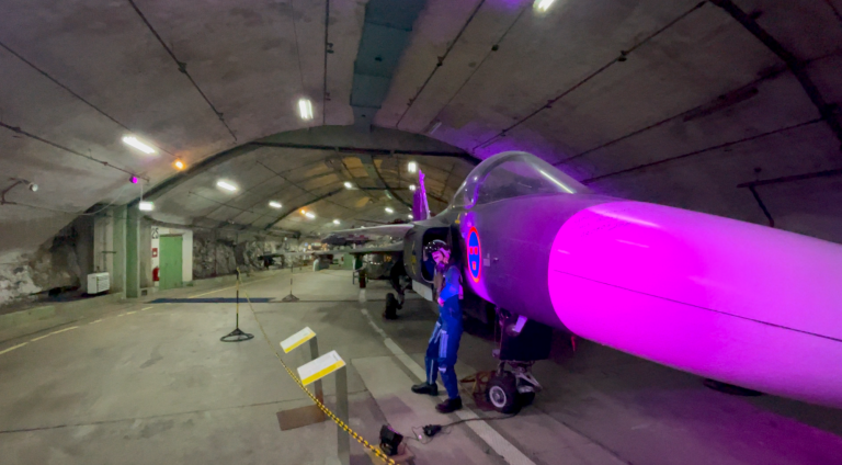 En pilot i blå dräkt står vid en stridsflygplans nos med lila belysning i ett hangar.