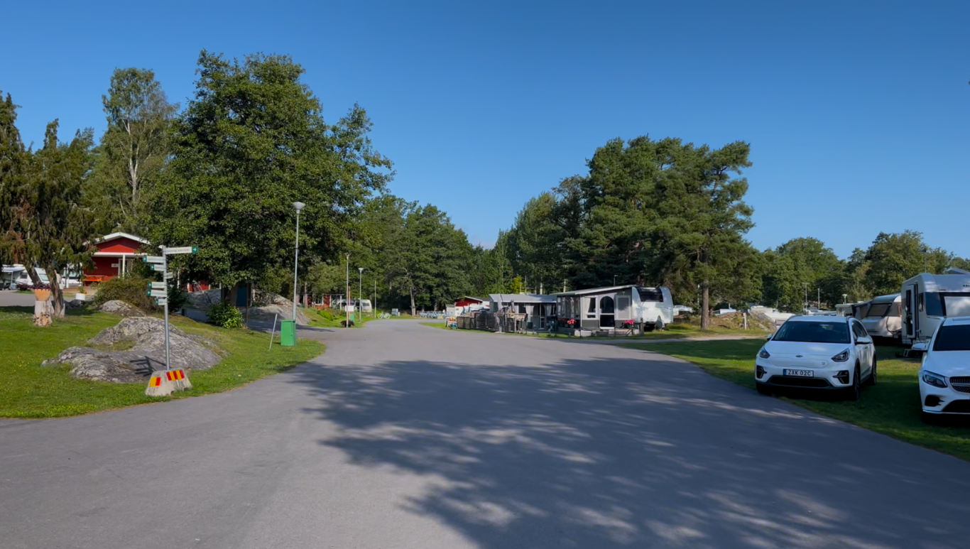 Incheckningen och bommen i horisonten En vid utsikt över en grusväg omgiven av träd och campingfordon.