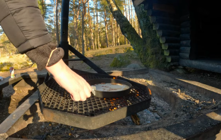 En hand placerar en panna på en eldstadsgrill i en skogsmiljö.