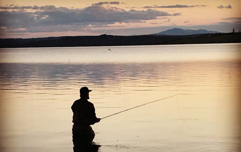 Person som fiskar vid vattenkant vid solnedgång. Berg i bakgrunden.