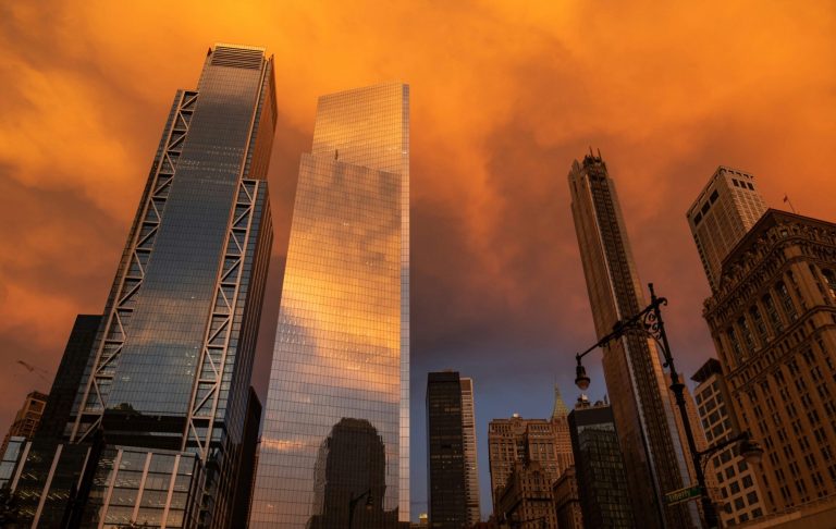 Höga skyskrapor reflekterar orange himmel vid solnedgång.