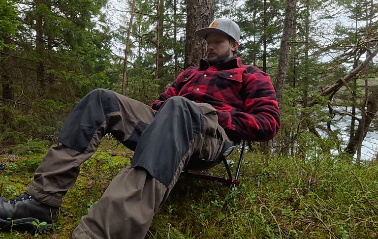 En person i rutig jacka sitter på en campingstol i skogsområde vid en sjö.