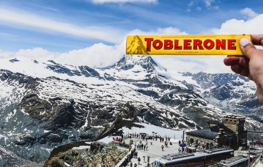 En hand håller en Toblerone framför matterhorn snötäckta fjäll och turister.