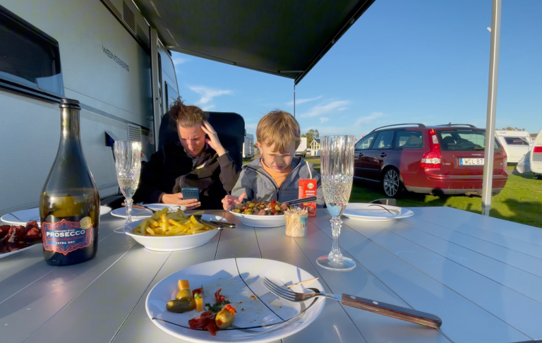 Två personer sitter vid ett bord utomhus, äter grilladmat och har drycker framför sig.